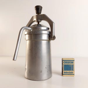 Puede incluir: Cafetera espresso plateada vintage con pomo y asa de madera. Una pequeña caja de cerillas "ORION televízió" está junto a la cafetera. La cafetera espresso tiene una palanca y un cuerpo cilíndrico.