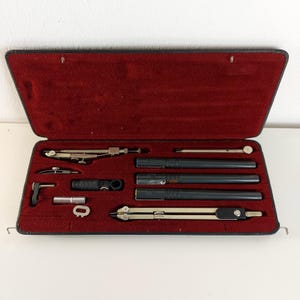 Könnte beinhalten: Ein offener schwarzer Koffer mit rotem Samtinnenfutter enthält ein Set von Zeichenwerkzeugen. Das Set umfasst mehrere schwarze Stifte, einen Zirkel und andere silberne und schwarze Instrumente. Der Koffer ist rechteckig und hat einen Klappdeckel.