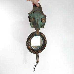 May include: A round leather-framed mirror with a brown leather strap hanging from the top. The mirror is surrounded by a braided leather design and has a small leather tassel hanging from the bottom.