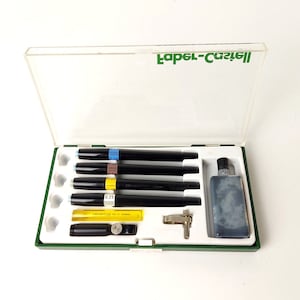 Op de afbeelding: Een transparante kunststof koffer met een groen deksel, met daarin een set van vijf zwarte technische pennen, een flesje zwarte inkt en een klein metalen gereedschap. Op het deksel staat de tekst "Faber-Castell" gedrukt.