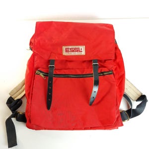 May include: A bright red backpack with black leather straps and a zippered front pocket. The backpack has a flap closure and adjustable shoulder straps. The brand name "ESSL" is displayed on a rectangular patch.
