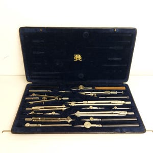 May include: A vintage drafting set in a dark blue velvet-lined case. The set includes various compasses, dividers, and other precision drawing tools. The case has a gold emblem on the lid and is open, displaying the tools neatly arranged.