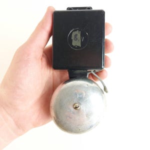 Può includere: Campana vintage nera e argentata con parte superiore quadrata e campana rotonda. La campana ha un piccolo numero 6 sbiadito. La campana è in metallo e ha un design classico.
