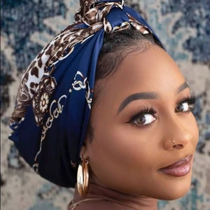 May include: A woman wearing a navy blue headscarf with a brown and white animal print and gold chain design. The headscarf is tied in a knot on top of the head. The woman is also wearing gold hoop earrings.
