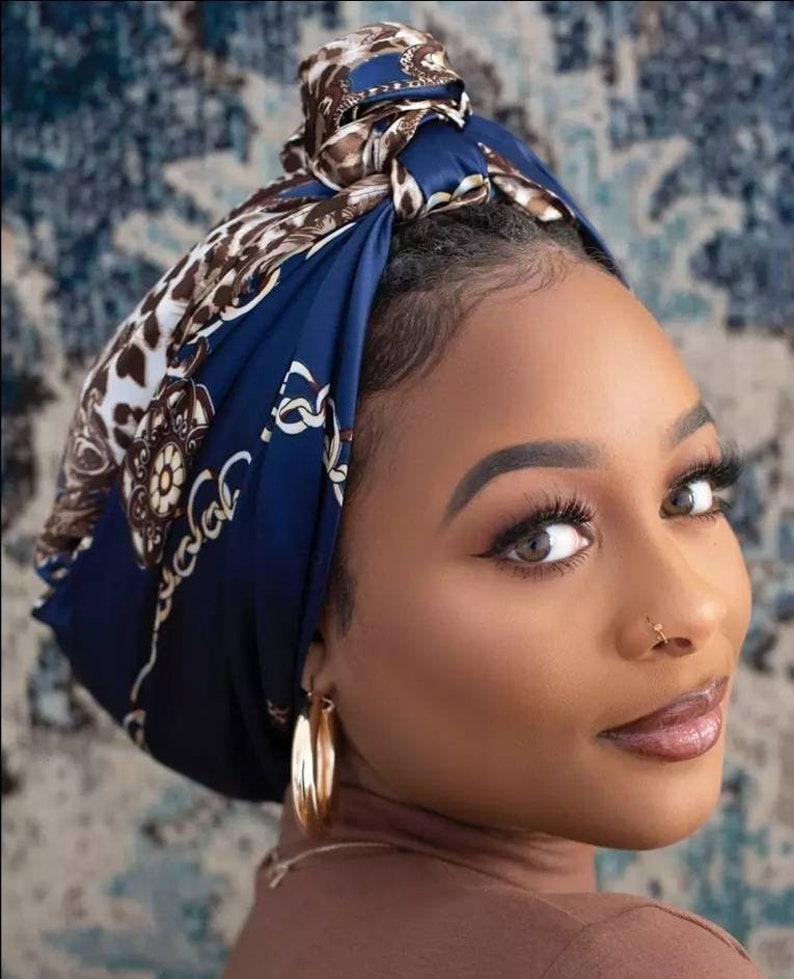 May include: A woman wearing a navy blue headscarf with a brown and white animal print and gold chain design. The headscarf is tied in a knot on top of the head. The woman is also wearing gold hoop earrings.