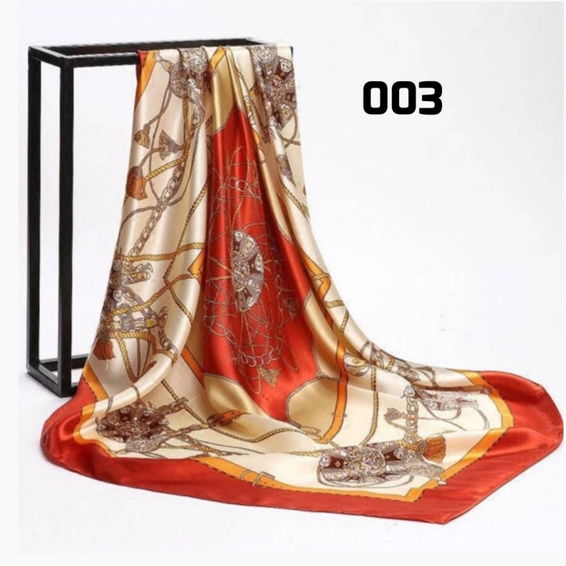 May include: A square silk scarf with an equestrian design. The scarf has a cream background with orange and brown details. The border is a solid orange, and the number "003" is printed on the scarf. The scarf is draped over a black stand.