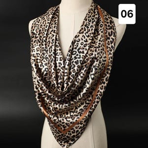 May include: A leopard print scarf draped over a mannequin. The scarf has a brown and black animal print pattern with a brown border. The scarf is made of a silky material and is displayed against a dark background.