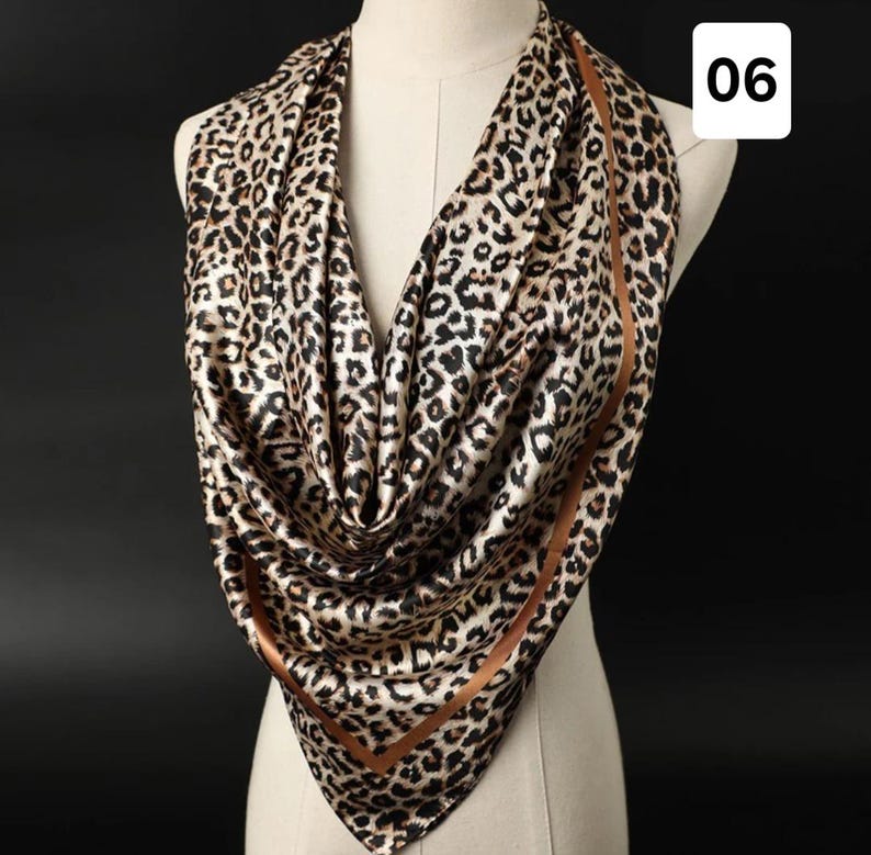 May include: A leopard print scarf draped over a mannequin. The scarf has a brown and black animal print pattern with a brown border. The scarf is made of a silky material and is displayed against a dark background.
