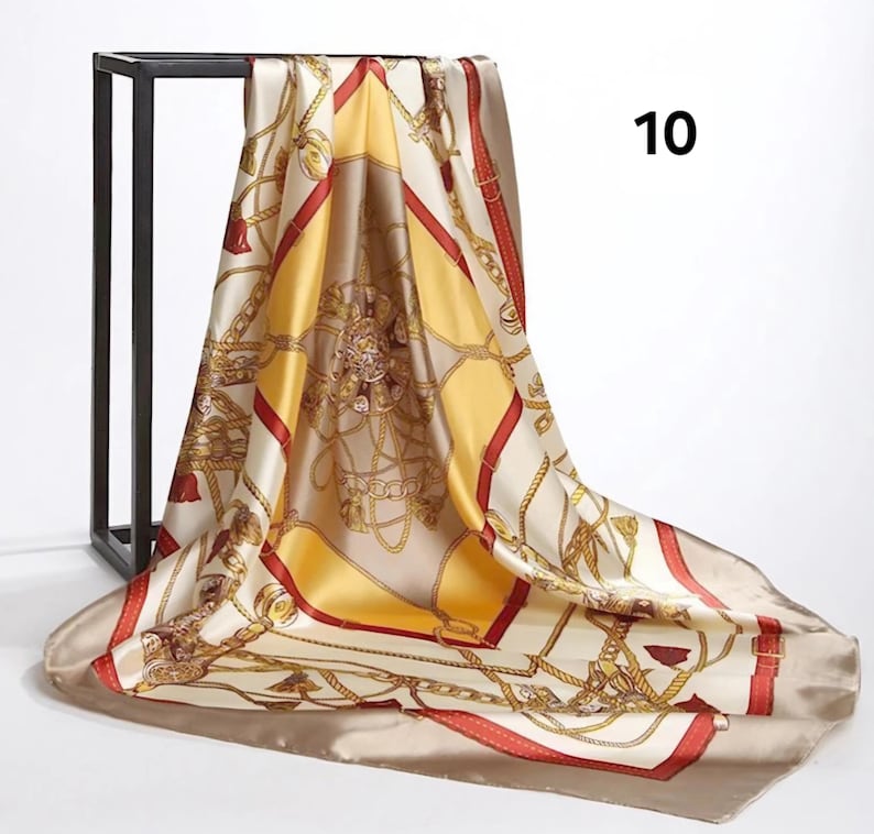 May include: A square silk scarf with a gold chain and buckle pattern on a cream background. The scarf has red and yellow accents and the number 10 in black in the upper right corner. The scarf is draped over a black metal stand.