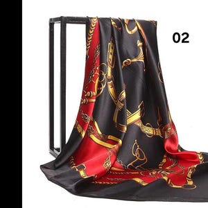 May include: A black and red silk scarf featuring a gold equestrian design. The scarf is displayed on a black metal stand. The number "02" is printed on the upper right. The scarf has a shiny, smooth texture and is made of silk.
