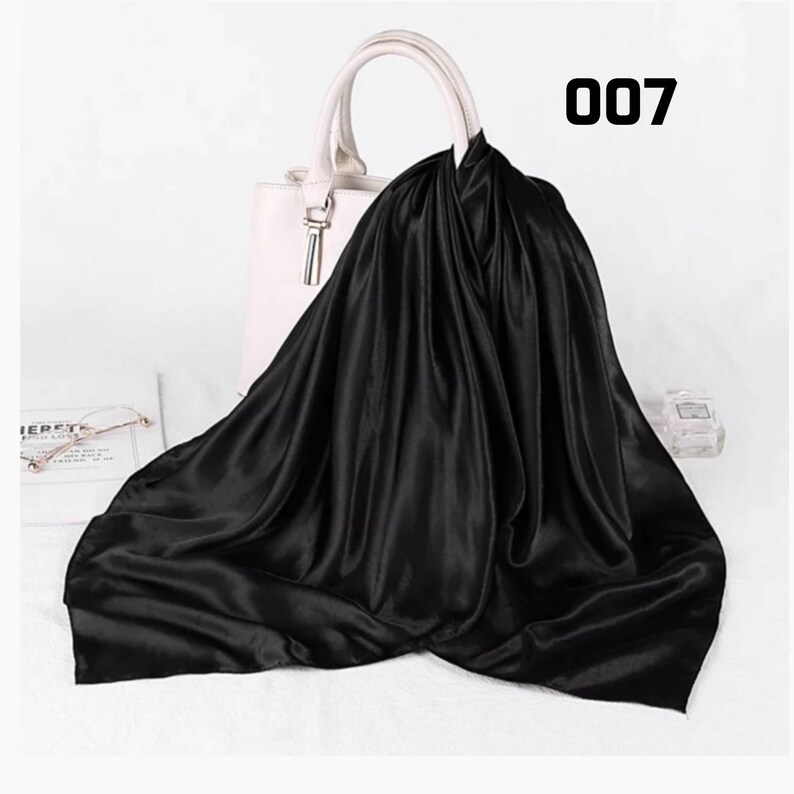 May include: A black satin scarf draped over a white handbag. The scarf has a glossy sheen and is folded to create soft, flowing lines. The number "007" is printed in black on the upper right corner. A pair of glasses and a perfume bottle are visible.