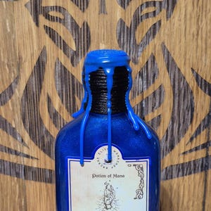 May include: A blue glass bottle with a black stopper and a label that reads "Potion of Mana". The label has a hand with a star above it and text that reads "Like a river of starlight, it rekindles spent energy, mends the frayed threads of magic, and awakens the boundless power lingering just beyond mortal grasp."