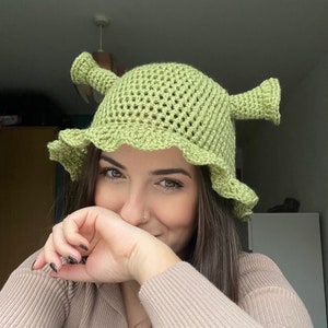 May include: A green crocheted hat with ears, resembling a fictional character. The hat is worn by a person with long brown hair, who is smiling and covering their face with their hand.