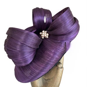May include: A deep purple fascinator hat featuring a structured, wide-brimmed base and a large, multi-looped bow made of stiffened, ribbed material. A cluster of small, round, cream-colored faux pearls is centered at the base of the bow loops.