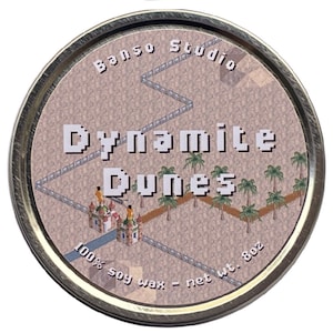 Puede incluir: Una lata dorada con una etiqueta beige que muestra una escena de desierto pixelada con palmeras, un río y un castillo. El texto "Dynamite Dunes" está impreso en letras grandes y pixeladas. La etiqueta también incluye el texto "100% soy wax - net wt. 8oz" y "Banso Studio".