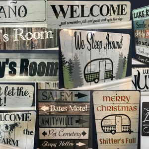 May include: A collection of rustic wooden signs with various text and designs. Signs include "El Ba&ntilde;o", "Welcome", "Zoey's Room", "Colby's Room", "Lake Rules", "We Sleep Around", and "Merry Christmas". Some signs feature illustrations.