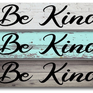 May include: Three wooden signs with the phrase "Be Kind" written in black cursive lettering. The signs have different wood grain patterns and colors, including white, light blue, and gray.