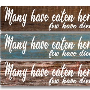 May include: Three rustic wooden signs with the text "Many have eaten here few have died" in white cursive font. The signs have different wood grain patterns and colors, including brown, blue, and gray.