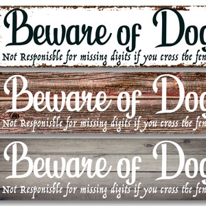 May include: Three wooden signs with the text "Beware of Dog" in white and black lettering. The signs have a rustic wood grain background and the text "Not Responsible for missing digits if you cross the fence" in black lettering.