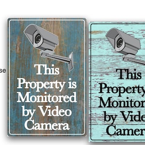 May include: Two rectangular signs with a distressed wood background and a security camera graphic. The signs read "This Property is Monitored by Video Camera" in white text. One sign has a blue background, the other is teal.
