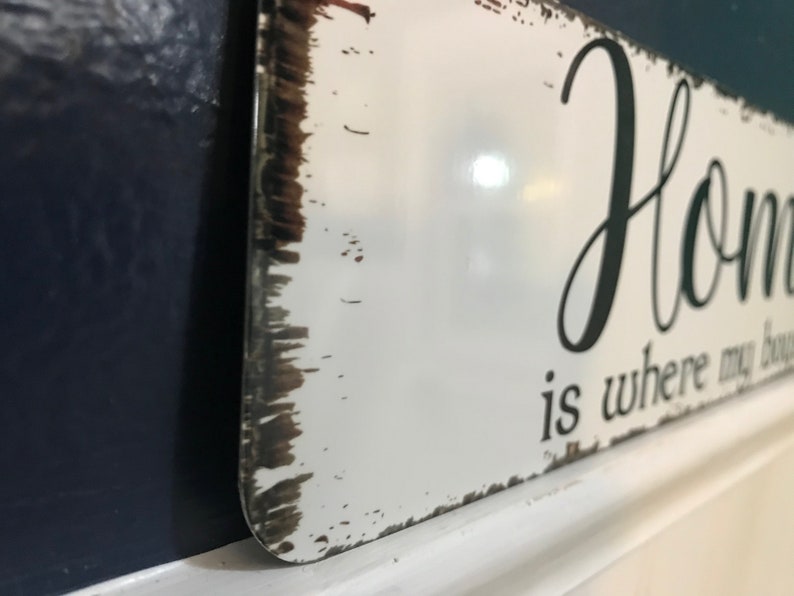 May include: White metal sign with the words "Home is where my bouquet is" in black script. The sign has a distressed, rustic look with brown edges and is mounted on a white surface against a dark blue wall.