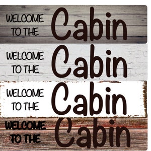 May include: Four wooden signs with the text "Welcome to the Cabin" in black lettering. The signs have different wood grain patterns and colors, including gray, white, and brown.