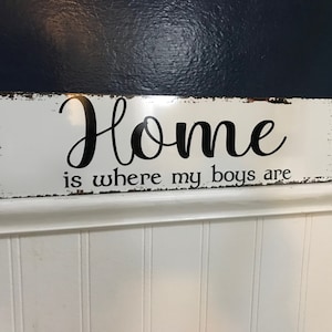 May include: A rectangular, distressed white sign with the phrase "Home is where my boys are" in black script. The sign has a rustic, vintage aesthetic, with visible wear and tear around the edges.