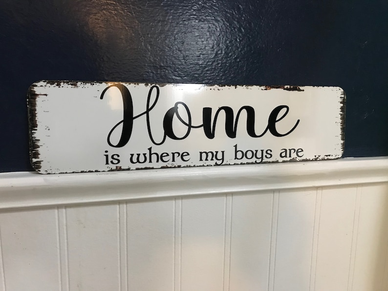 May include: A rectangular, distressed white sign with the phrase "Home is where my boys are" in black script. The sign has a rustic, vintage aesthetic, with visible wear and tear around the edges.