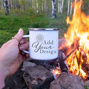 May include: White enamel camping mug with a black mandala design and the text "Add Your Design". The mug is being held by a hand in front of a campfire.