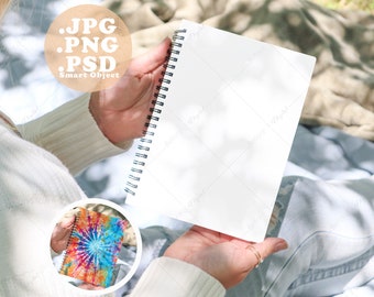 May include: A person holding a blank white spiral notebook with a black spiral binding. The notebook is sitting on a person's lap. The person is wearing a white sweater and blue jeans.
