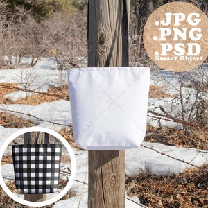 May include: A white tote bag mockup is displayed on a wooden post, showcasing a blank canvas for design. A circular inset reveals a black and white checkered pattern. The background features a natural outdoor setting with barbed wire and winter foliage.