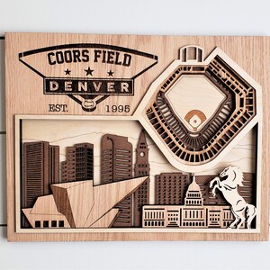 May include: A wooden wall art piece depicting Coors Field, a baseball stadium in Denver, Colorado. The art features a 3D layered design of the stadium, the Denver skyline, and a rearing horse. The text "Coors Field Denver Est. 1995" is engraved on the top of the piece.