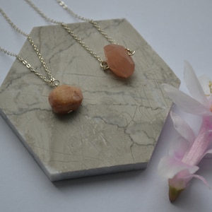 Könnte beinhalten: Zwei zarte silberne Halsketten mit rosafarbenen Edelsteinanhängern auf einem Marmor-Sechseck. Die Halsketten werden auf einem weißen Hintergrund mit einer rosa Blume präsentiert.