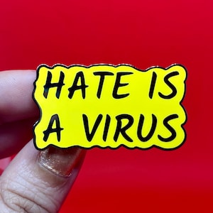 Puede incluir: Un pin de esmalte amarillo con las palabras "HATE IS A VIRUS" en letras negras y en negrita. El pin tiene un borde ondulado e irregular y un contorno plateado. El fondo es rojo liso.