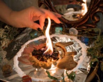 May include: A hand holding a lit stick over a small dish with a burning brown substance. The dish is surrounded by white salt and green herbs. There is a mirror in the background.
