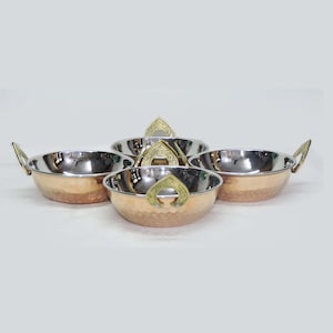 May include: Set of four copper-colored serving bowls with hammered exteriors and shiny silver interiors. Each bowl has two small, gold-colored handles. The bowls are arranged in a circular pattern, showcasing their design and craftsmanship.