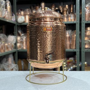 May include: A copper water dispenser with a hammered texture, a spigot, and a lid. The dispenser sits on a gold-colored metal stand. The "SAGA" logo is visible on the front of the dispenser. The dispenser is designed to hold a large volume of liquid.