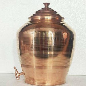 May include: A copper-coloured water dispenser with a lid and a spigot. The dispenser has a round body with horizontal etched lines and a decorative lid. The spigot is located near the bottom of the dispenser.