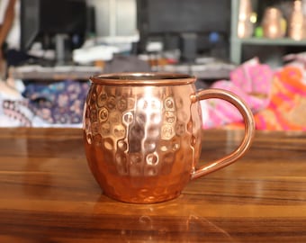 May include: A copper Moscow Mule mug with a hammered texture. The mug has a rounded body, a handle, and a polished finish. It sits on a wooden surface. The background is blurred.