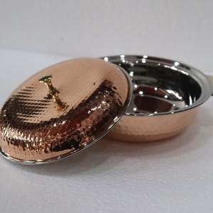 May include: A copper-colored serving dish with a hammered texture and a matching lid. The lid is slightly ajar, revealing the stainless steel interior of the dish. A small, gold-colored handle is on the lid, and a gold-colored handle is on the side of the dish.