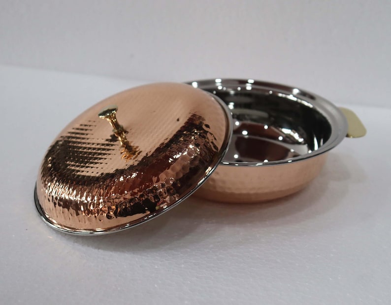 May include: A copper-colored serving dish with a hammered texture and a matching lid. The lid is slightly ajar, revealing the stainless steel interior of the dish. A small, gold-colored handle is on the lid, and a gold-colored handle is on the side of the dish.