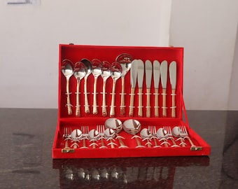 May include: A complete silver-colored flatware set with gold-colored handles, displayed in a red velvet-lined case. The set includes spoons, forks, knives, and serving spoons. The case is open, showcasing the arrangement of the utensils.