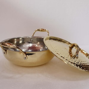 Op de afbeelding: Een goudkleurige metalen kookpot met bijpassende deksel. De pot heeft twee handvatten en een gehamerde textuur aan de binnenkant. Het deksel is gedeeltelijk open en onthult de binnenkant van de pot.