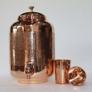 May include: A copper water dispenser with a lid, spigot, and two matching cups. The dispenser has an etched floral design. The cups and dispenser are made of shiny copper.