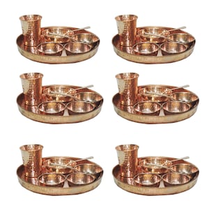 May include: Six copper-colored thali sets, each with a hammered texture. Each set includes a large oval tray, a tall glass, small bowls, and spoons. The sets are arranged in a grid pattern, showcasing the complete dining sets.