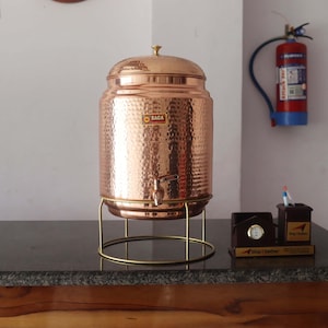 May include: A hammered copper water dispenser with a spigot, lid, and a gold-colored stand. The dispenser has the word "SAGA" on the front. The dispenser is on a dark countertop.