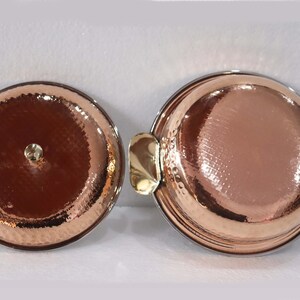 May include: Two copper-colored serving dishes with hammered textures. One has a lid with a small gold-colored knob. The other has two gold-colored handles. The dishes are round and have a shiny finish.