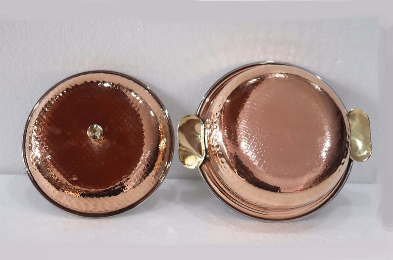 May include: Two copper-colored serving dishes with hammered textures. One has a lid with a small gold-colored knob. The other has two gold-colored handles. The dishes are round and have a shiny finish.