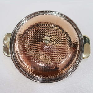 May include: A copper-colored serving dish with a hammered texture, a silver-colored rim, and gold-colored handles. The lid has a small gold-colored knob. The dish is round and appears to be made of metal.