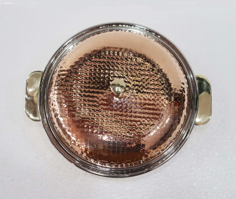 May include: A copper-colored serving dish with a hammered texture, a silver-colored rim, and gold-colored handles. The lid has a small gold-colored knob. The dish is round and appears to be made of metal.